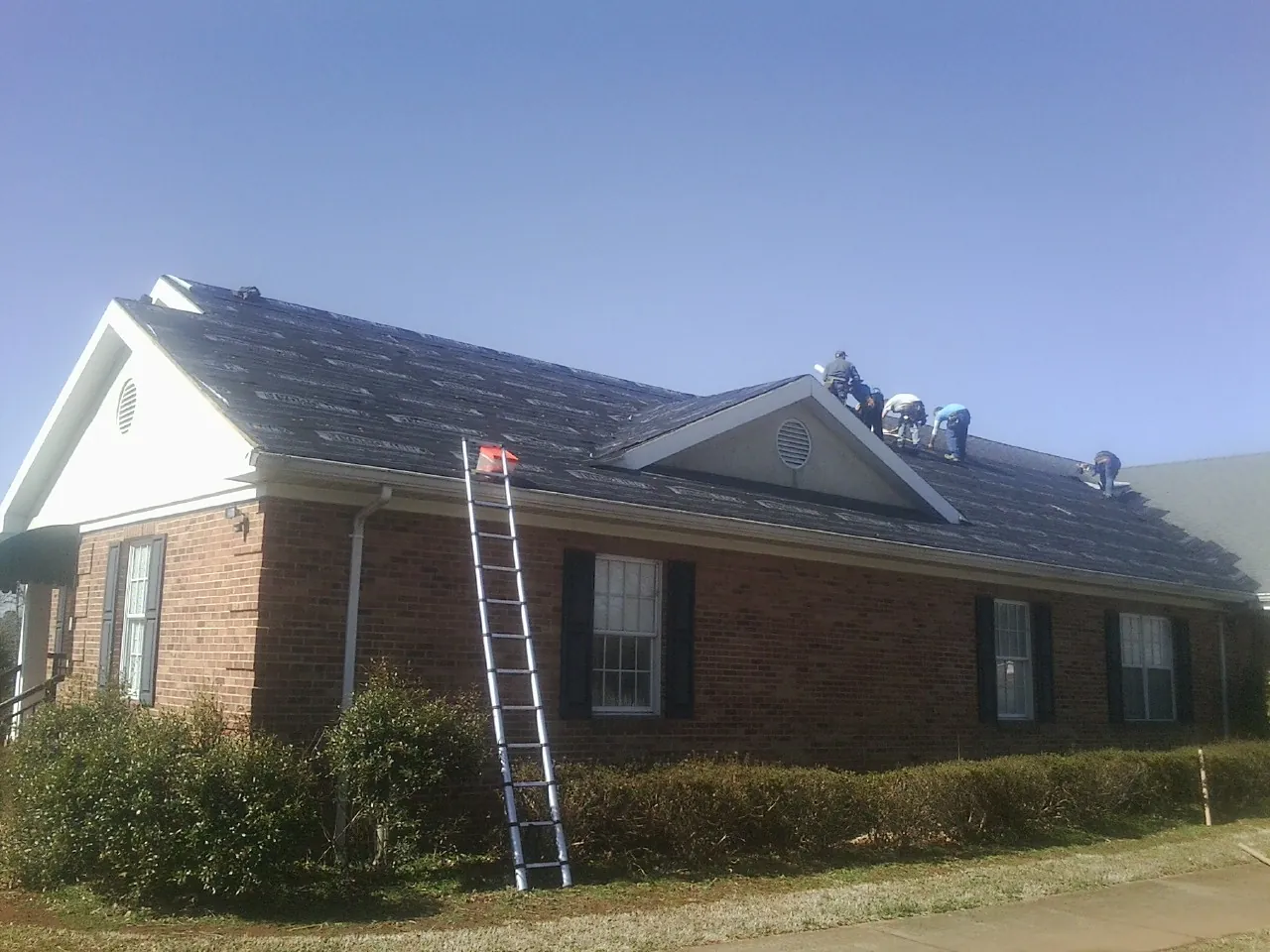 Crew tearing shingles off a brick ranch roof with a ladder set — Emergency Roof Repair work by our crew in Maben