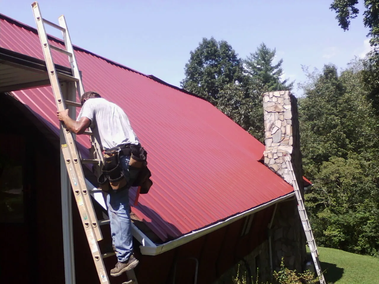 Roofer on a ladder beside a red metal roof and stone chimney — Soffit & Fascia Repair by Roofing Brothers in Maben